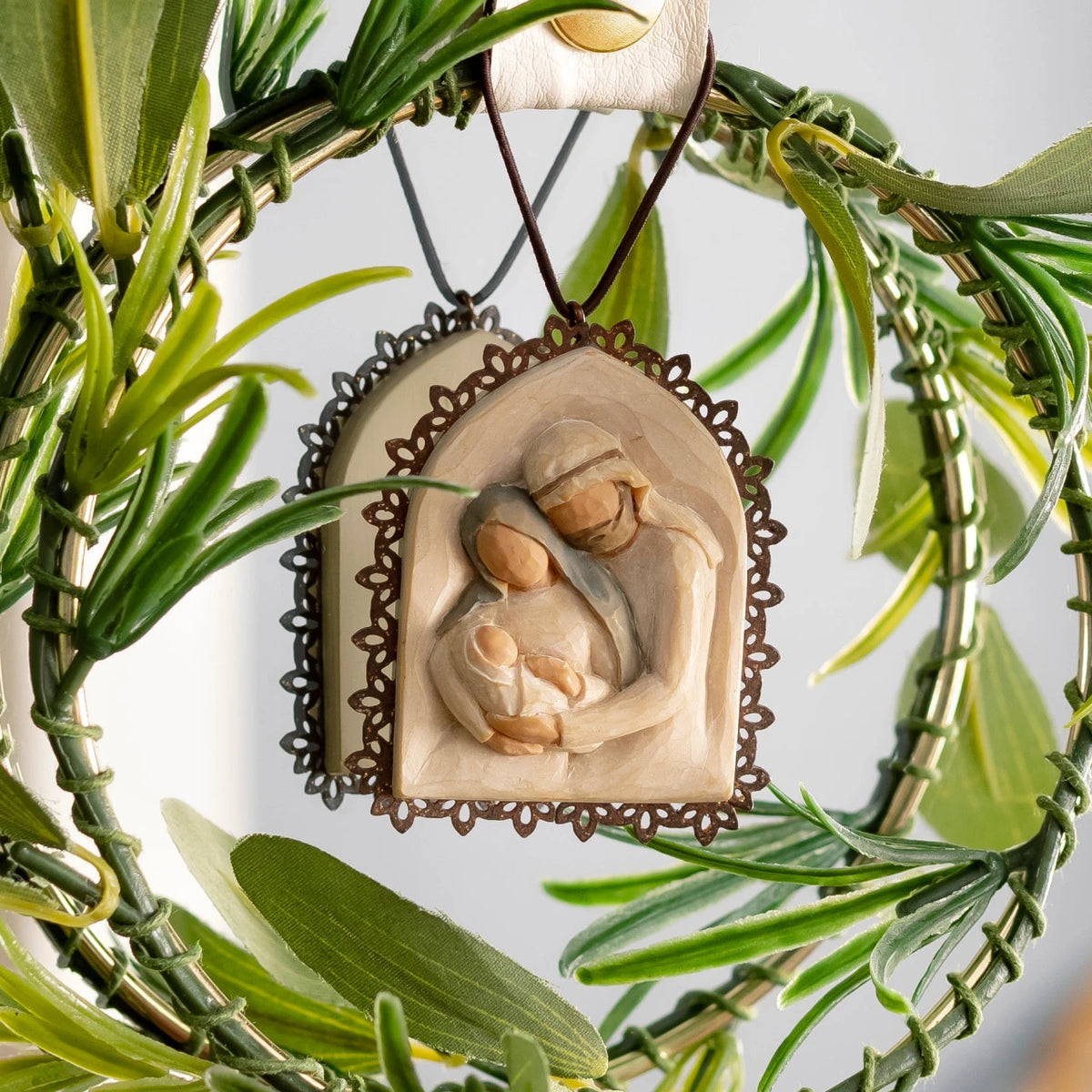 Willow Tree Holy Family Metal-edged Ornament, 2”w x 2.25”h hand-painted resin bas-relief carving of Joseph and Mary holding baby Jesus, edged in pierced-metal filigree, brown elastic cord, hanging in greenery wreath