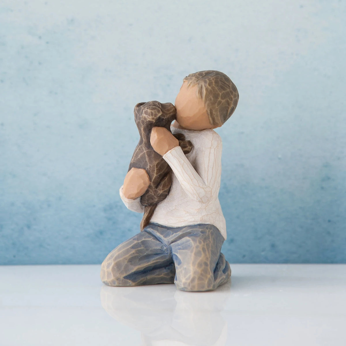 Young faceless boy figurine wearing white shirt and blue jeans sitting holding brown puppy - background is blue