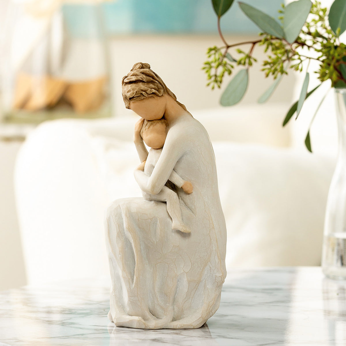 Willow Tree For Always, 6.5”h hand-painted resin female figure, seated, holding toddler on lap, both in cream clothes, arms around each other, on table with florals