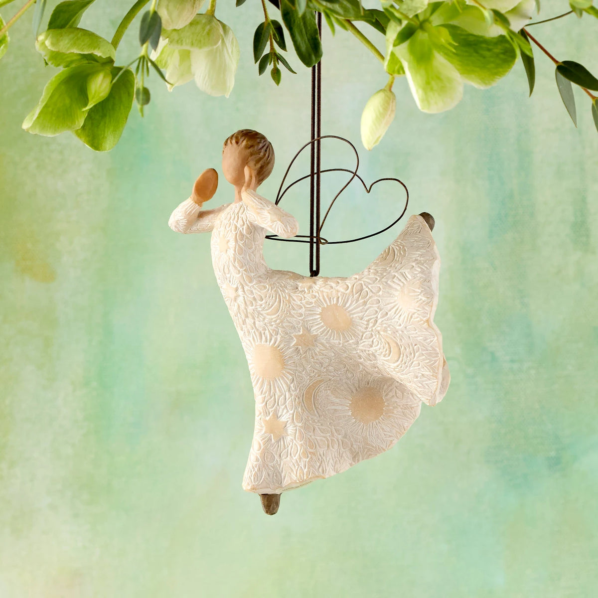 Willow Tree Song of Joy Ornament, 5”h hand-painted resin angel figure in ballet pose with hands raised to mouth, in cream dress carved with celestial symbols, with wire wings, brown elastic cord, hanging in front of green painting