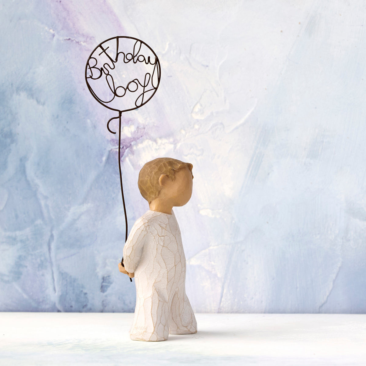 Willow Tree Birthday Boy, 5”h hand-painted standing resin figure in cream onesie, holding wire ballon with 'birthday boy' written in wire, in front of blue painting