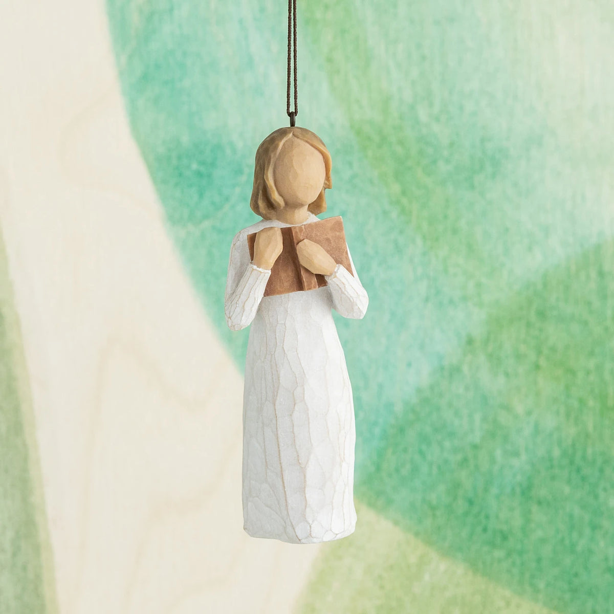 Willow Tree Love of Learning Ornament, 4”h hand-painted resin figure in cream dress holding open book to her chest, brown elastic cord, hanging in front of cyan painting