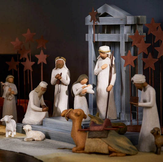 A carved nativity scene set out on a wood table laid with a linen cloth and lit candles in the background.