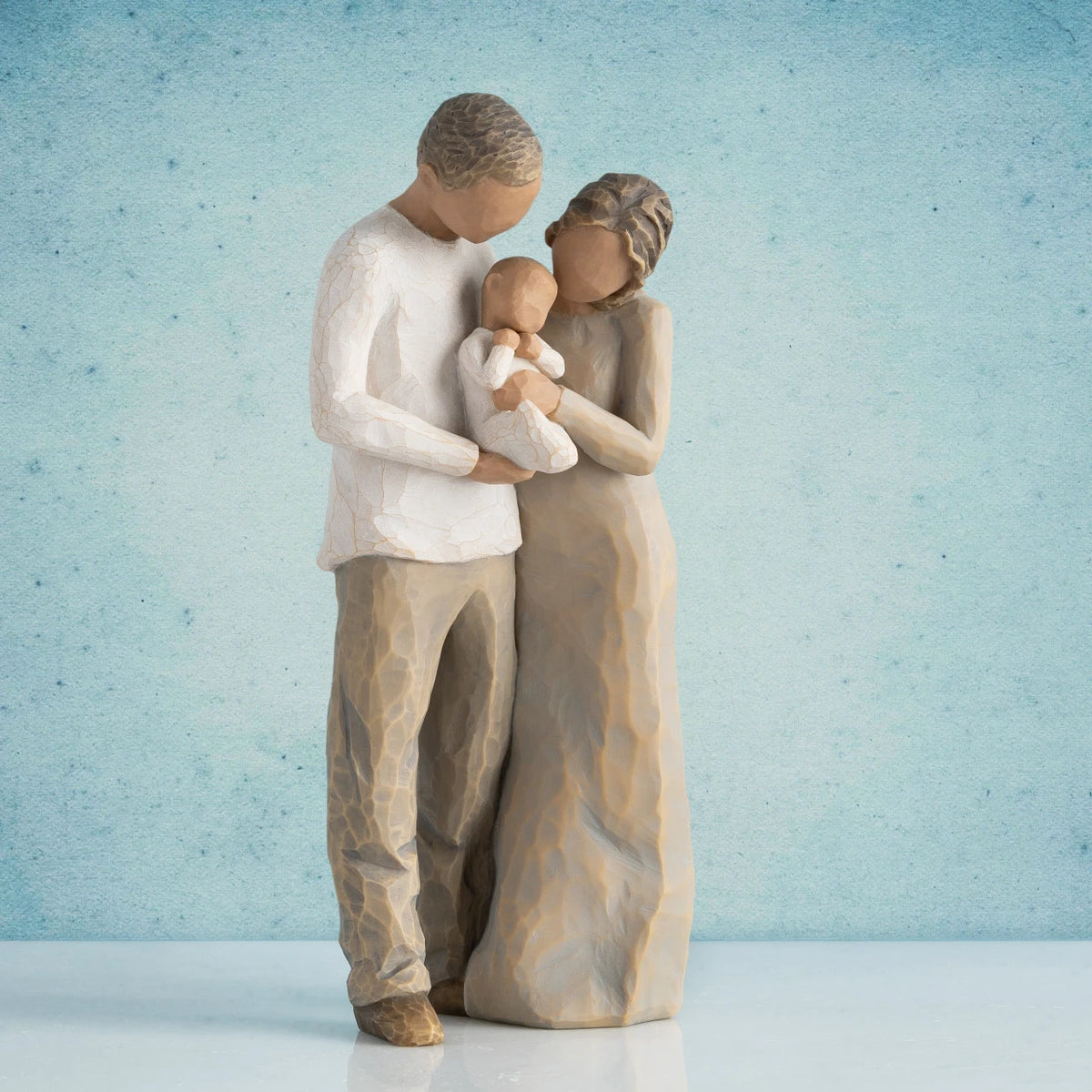 Willow Tree We are Three, 8.5”h hand-painted figure of couple standing together, holding baby between them, all in neutral clothing with darker skin and hair, in front of blue painting