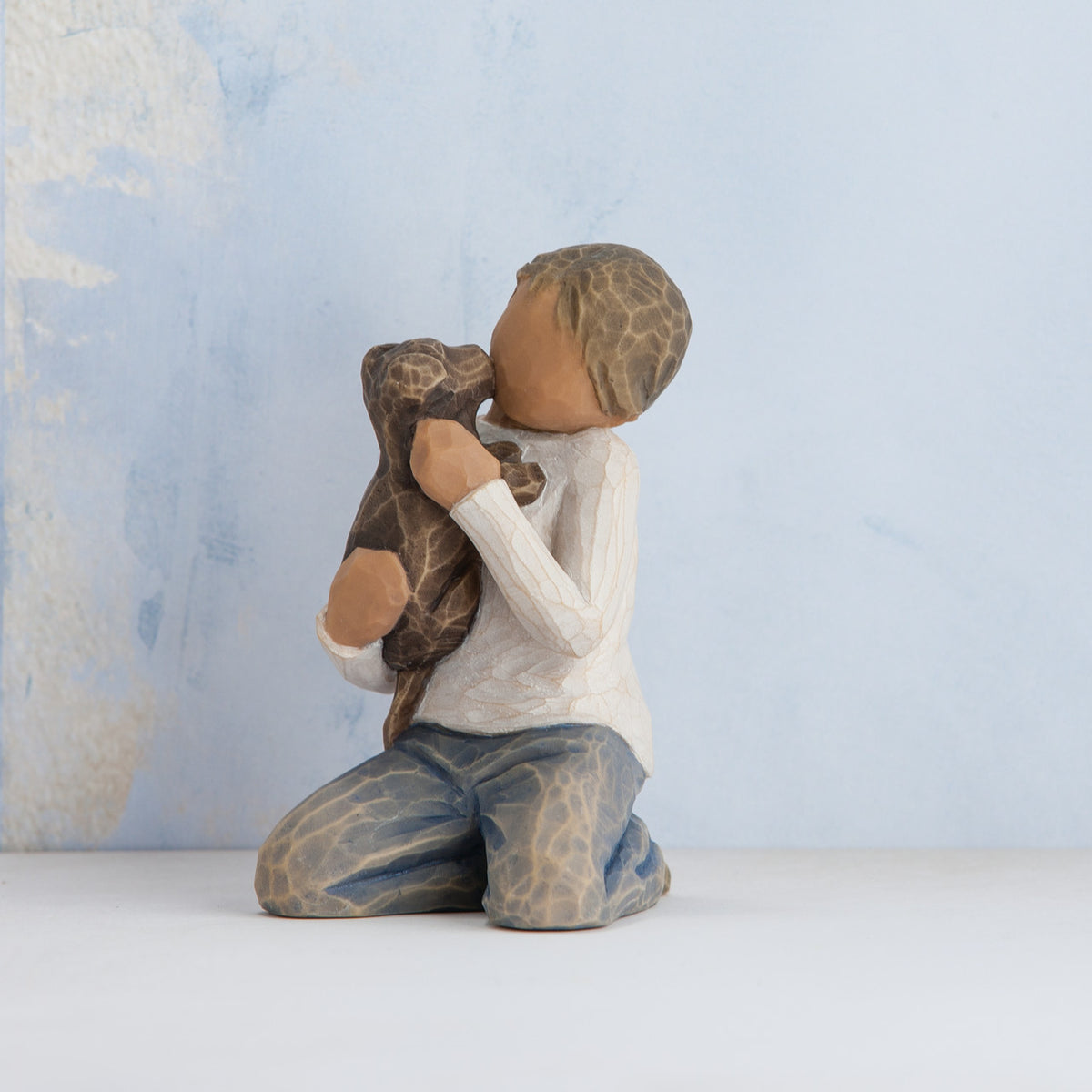 Willow Tree Kindness (boy), 3" hand-painted resin figure in neutral clothes, darker skin and hair, kneeling and holding black dog, in front of pale blue painting