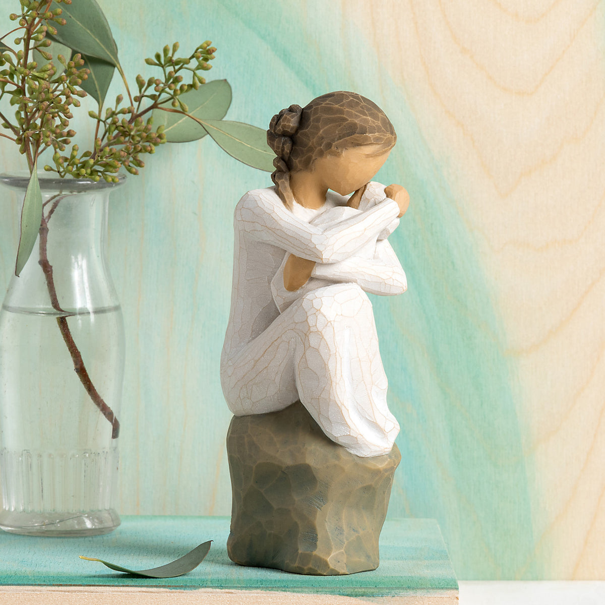 Willow Tree Guardian, 6”h hand-painted resin figure in cream dress, seated on gray rock, holding newborn baby in cream blanket, in front of celedon painting