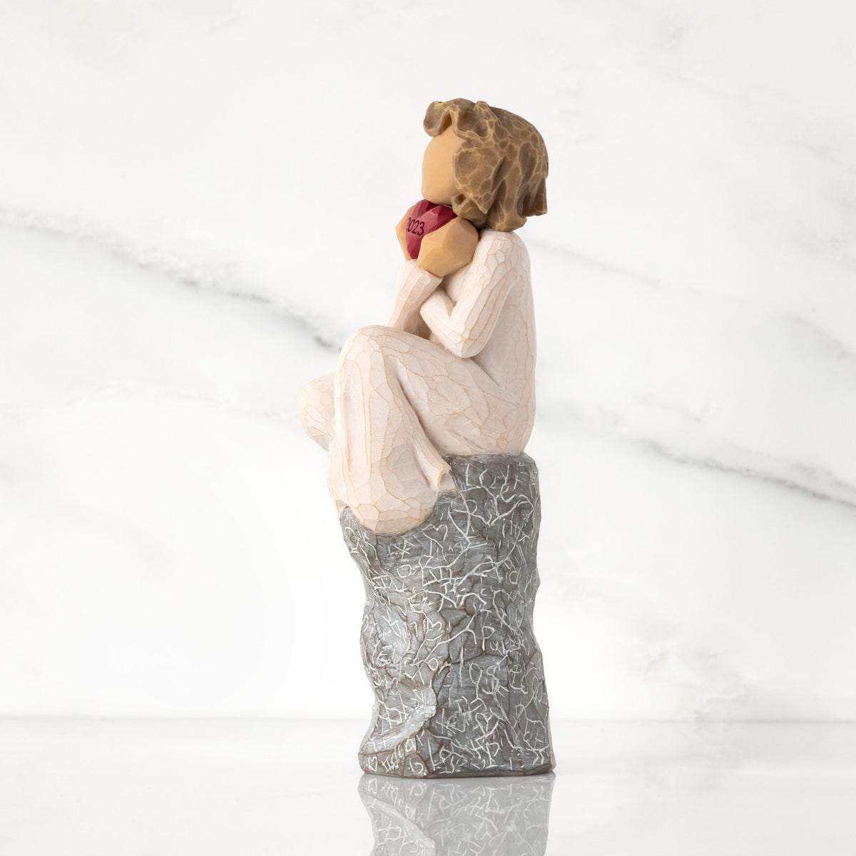Facing left: figure in cream dress, seated on gray rock, knees drawn up to chest, holding red heart engraved with year 2023