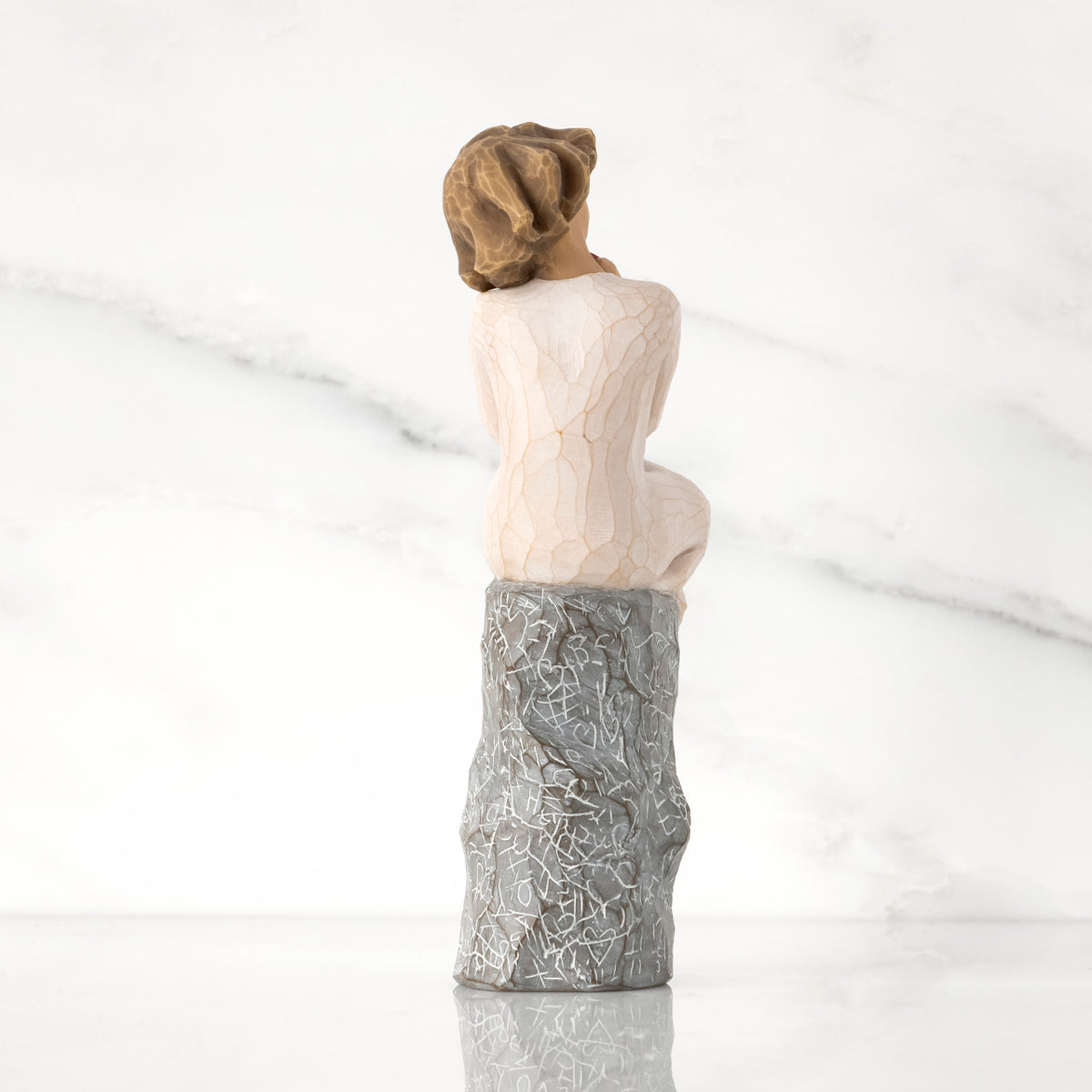 Back view: figure in cream dress, seated on gray rock, rock carved with symbols of love