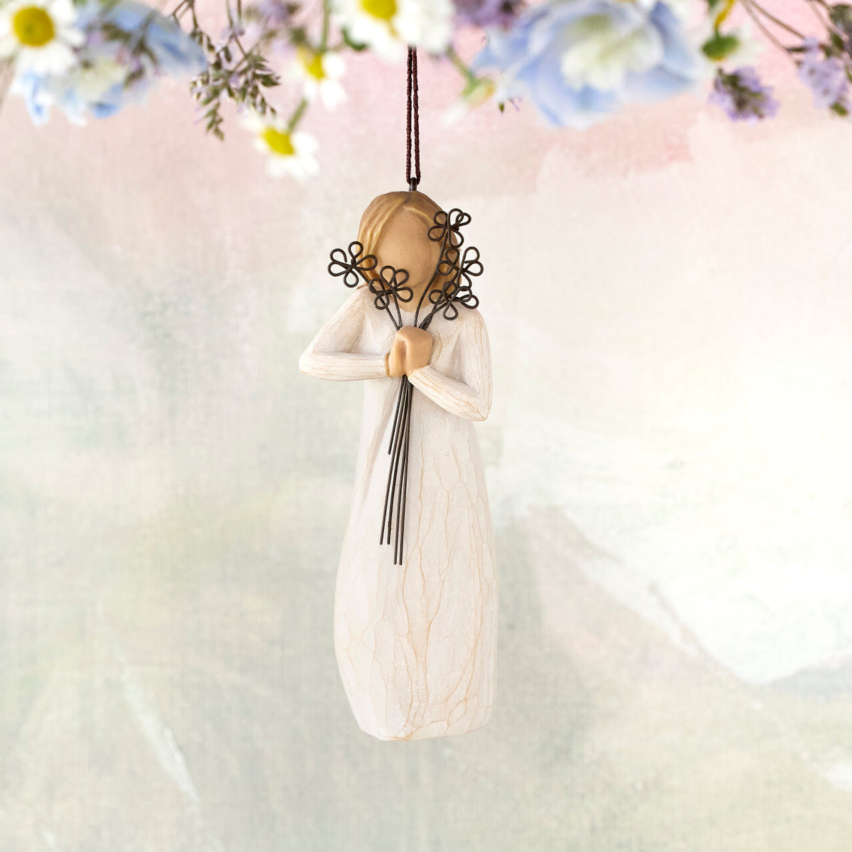 Willow Tree Friendship Ornament, 5.5"h hand-painted resin figure in cream dress holding wire flowers with long stems, brown elastic cord, hanging in front of pastel painting