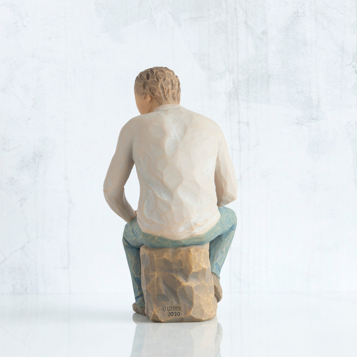 Back view, adult male figure in light gray shirt and blue jeans, seated on gray rock