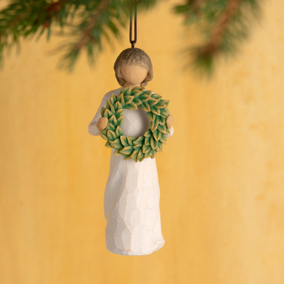 Willow Tree Magnolia Ornament, 4.25”h hand-painted resin figure in cream dress, holding green magnolia leaf wreath, brown elastic cord, hanging in front of light orange painting