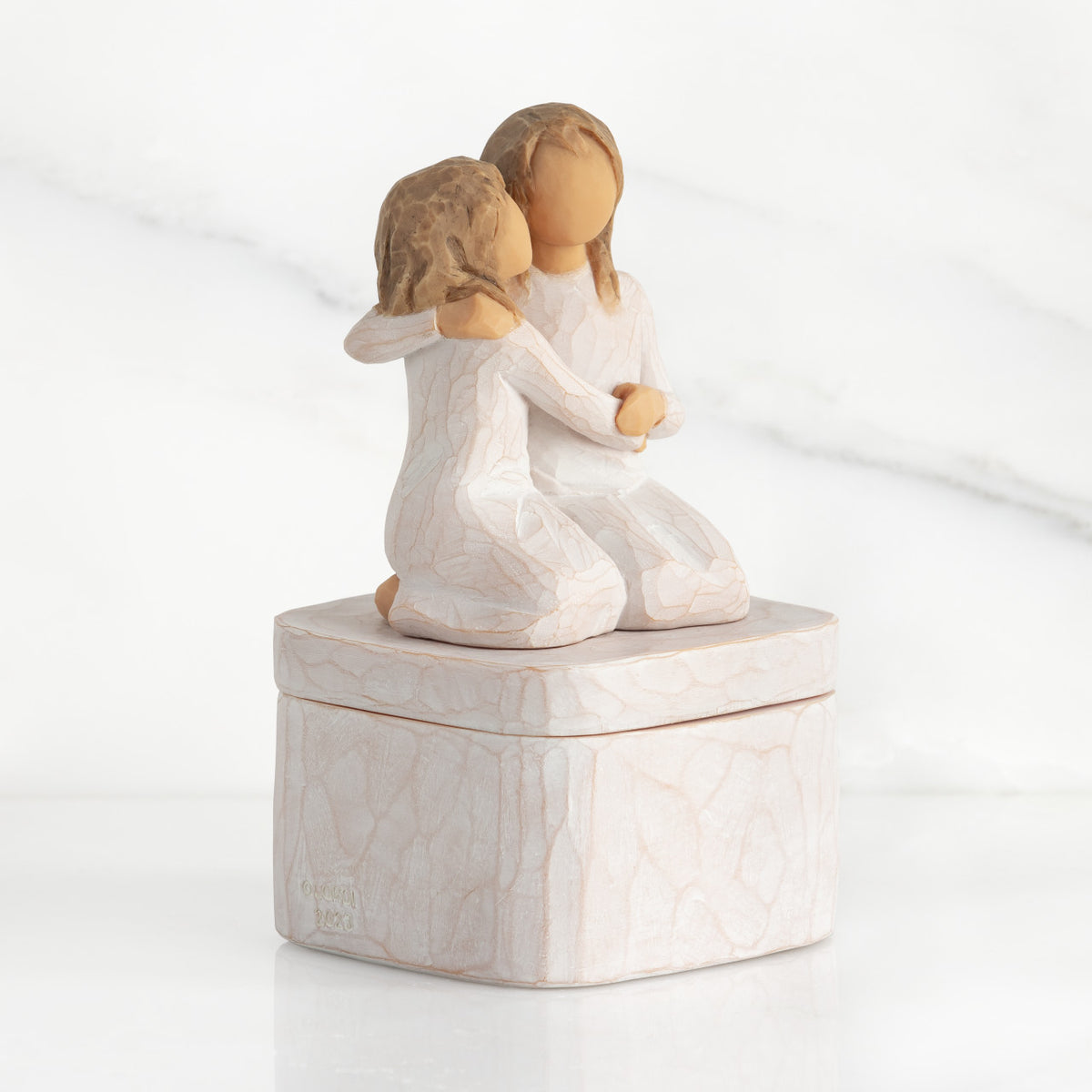 Right side: square resin box, two 3-D figures kneeling atop lid, in cream dresses, hugging each other