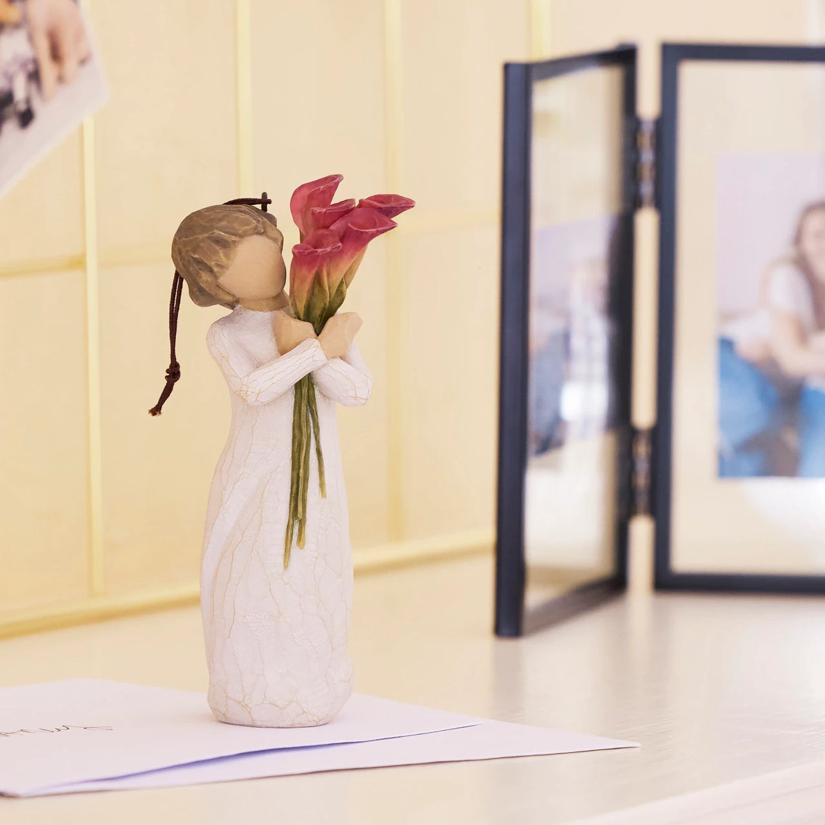 Willow Tree Bloom Ornament, 4.5”h hand-painted resin figure in cream dress, holding bouquet of fuchsia calla lilies, brown elastic cord, on desk next to photo