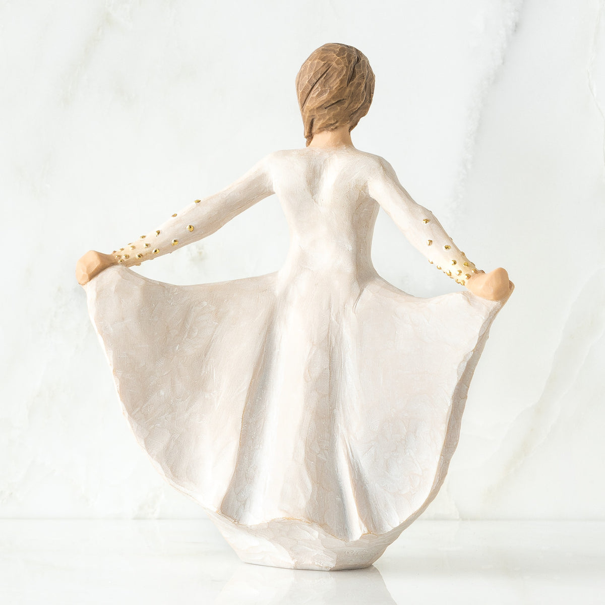 Back view of standing female figure in cream dress, holding out her skirt, which is carved with butterfly markings and dotted with gold