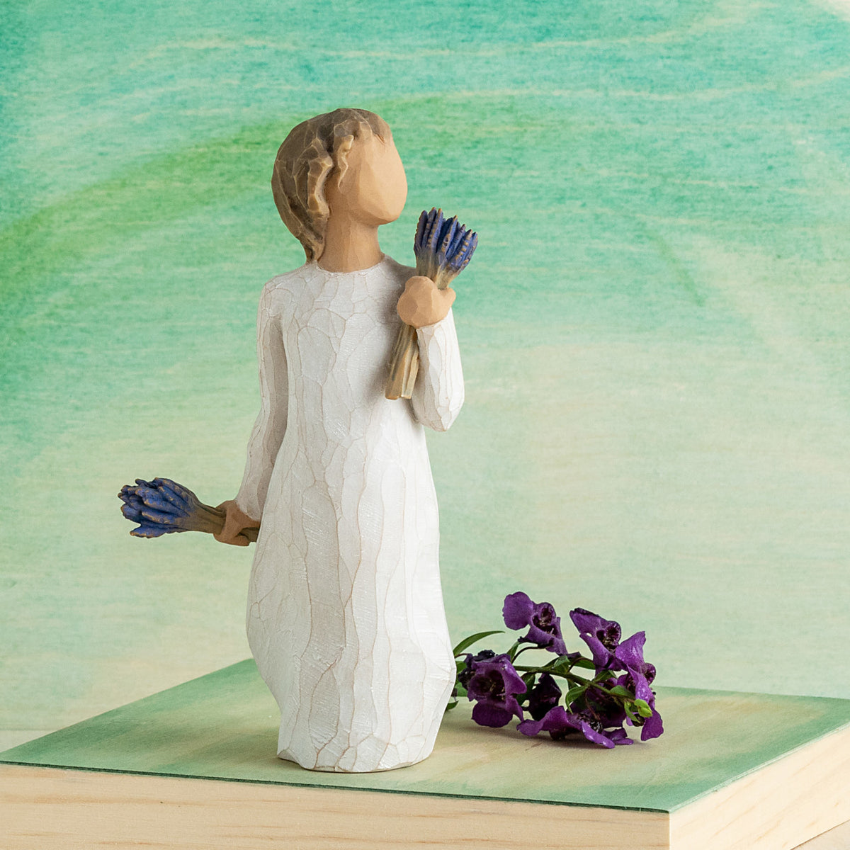 Willow Tree Lavender Grace, 5.5”h hand-painted standing resin figure in cream dress, holding two bundles of lavender flowers - one in raised hand, one at her side, in front of a teal painting