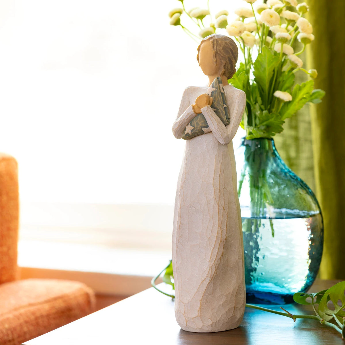 Willow Tree Hero, 9.5”h hand-painted standing resin figure in cream dress holding folded American flag in her arms, on table with vase of yellow flowers