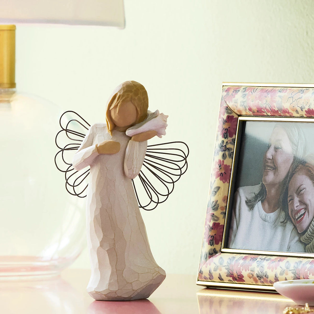 Willow Tree Thinking of You, 5.5”h hand-painted standing resin angel figure in cream dress with wire wings, holding pink conch shell to ear, on shelf with photos