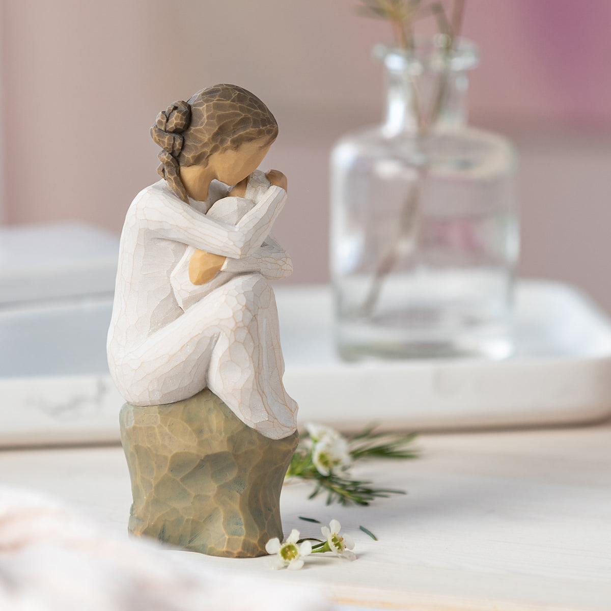 Willow Tree Guardian, 6”h hand-painted resin figure in cream dress, seated on gray rock, holding newborn baby in cream blanket, on table with glass vase