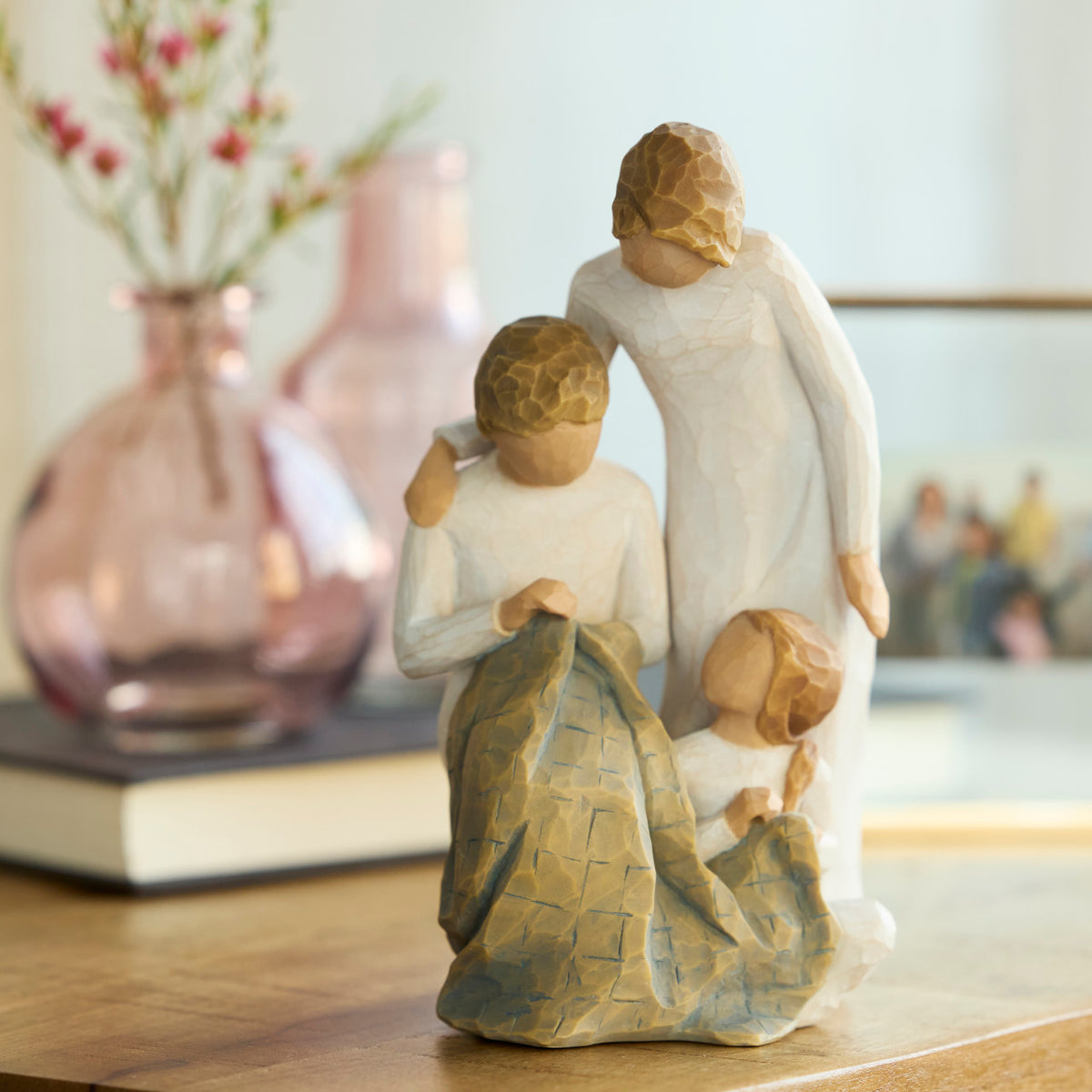 Willow Tree Generations, 7”h hand-painted resin figure of seated older woman sewing blue quilt on her lap; standing woman at her shoulder; seated young girl at her feet, all in neutral clothing, on table with pink glass vase