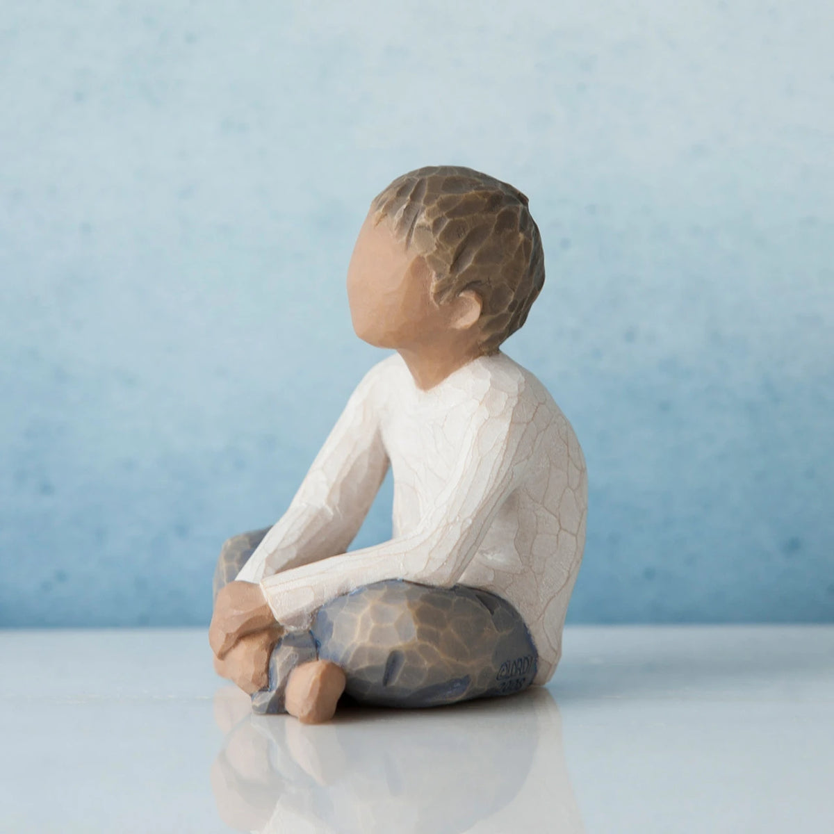 Small brunette boy figurine sitting to the side wearing white shirt and blue jeans and hands in lap - blue background