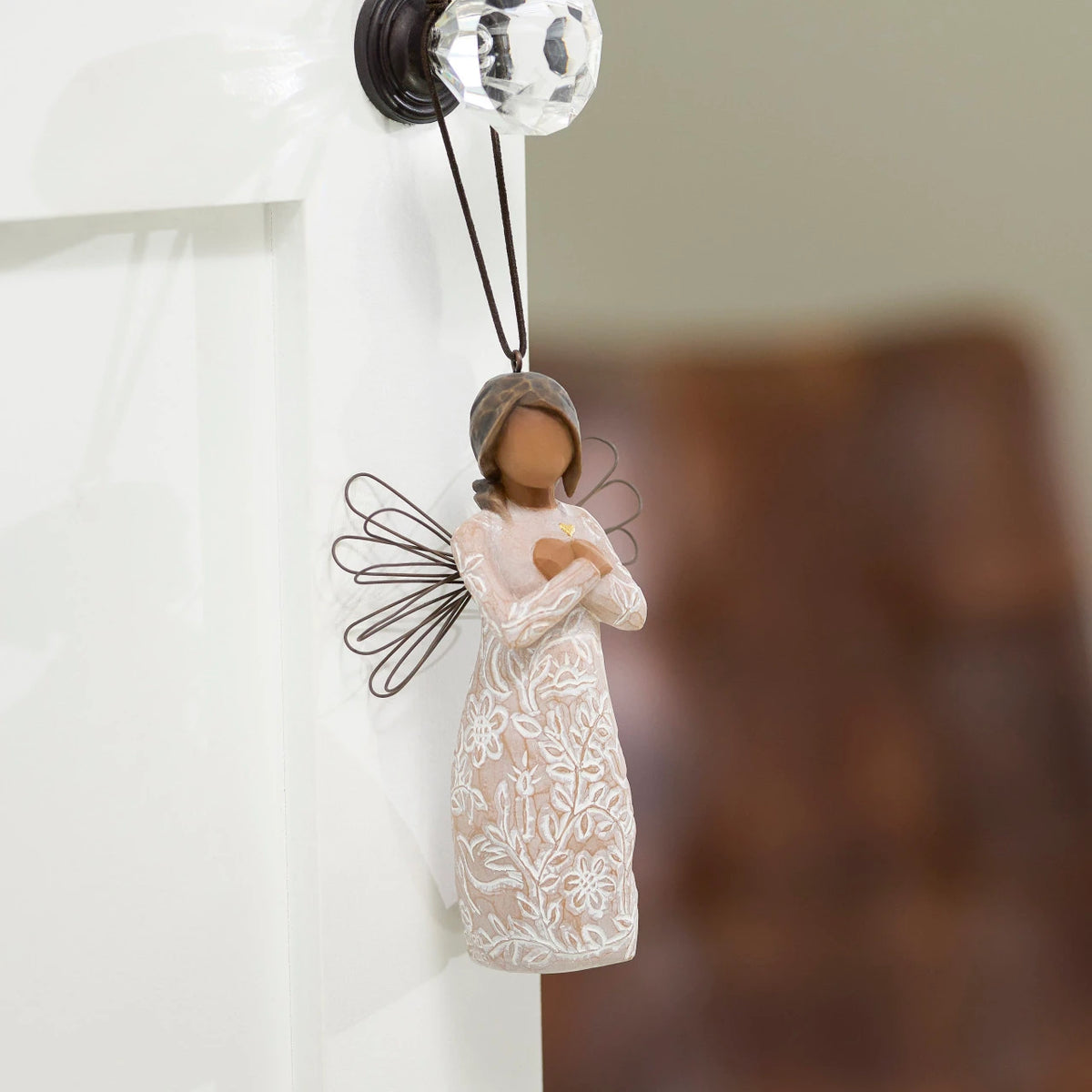Willow Tree Remembrance Ornament, 4”h hand-painted resin angel figure with darker skin and hair, wire wings, arms crossed over gold heart, cream dress carved with nature imagery, brown elastic cord, hanging on glass doorknob