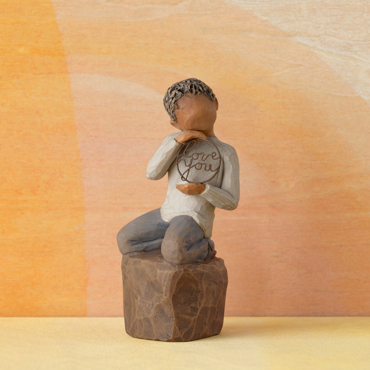 Willow Tree Love you too (darker skin), 4.5”h hand-painted resin figure of young child in cream shirt and jeans, kneeling on rock, holding wire circle with ‘love you’ inside, in front of orange painting