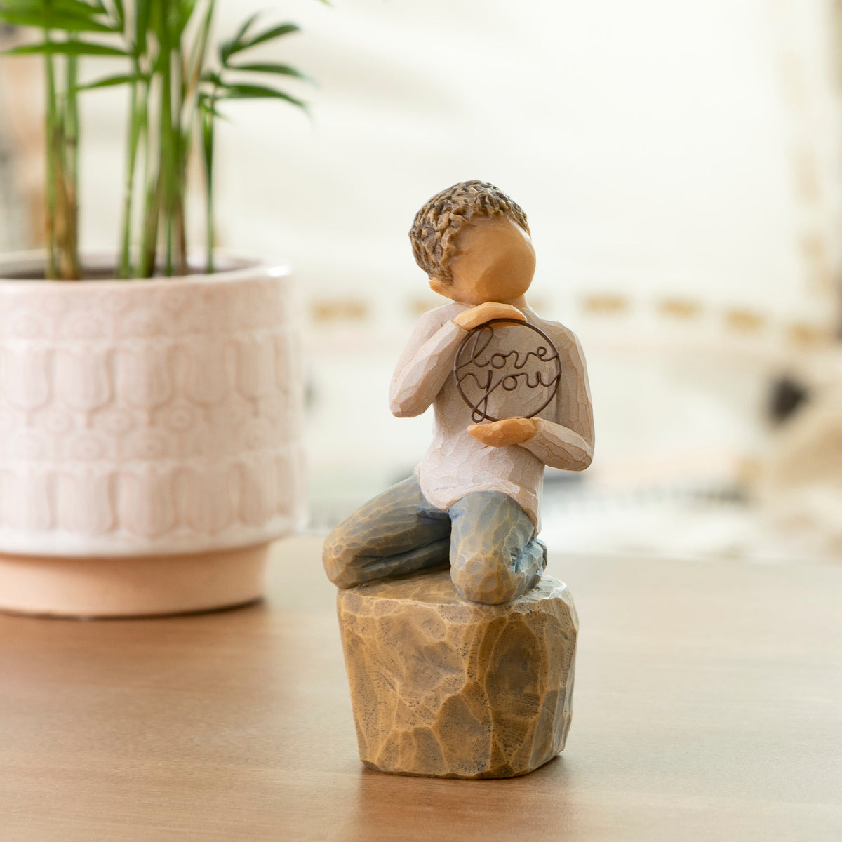 Willow Tree Love you too (lighter skin), 4.5”h hand-painted resin figure of young child in cream shirt and jeans, kneeling on rock, holding wire circle with ‘love you’ inside, on coffee table