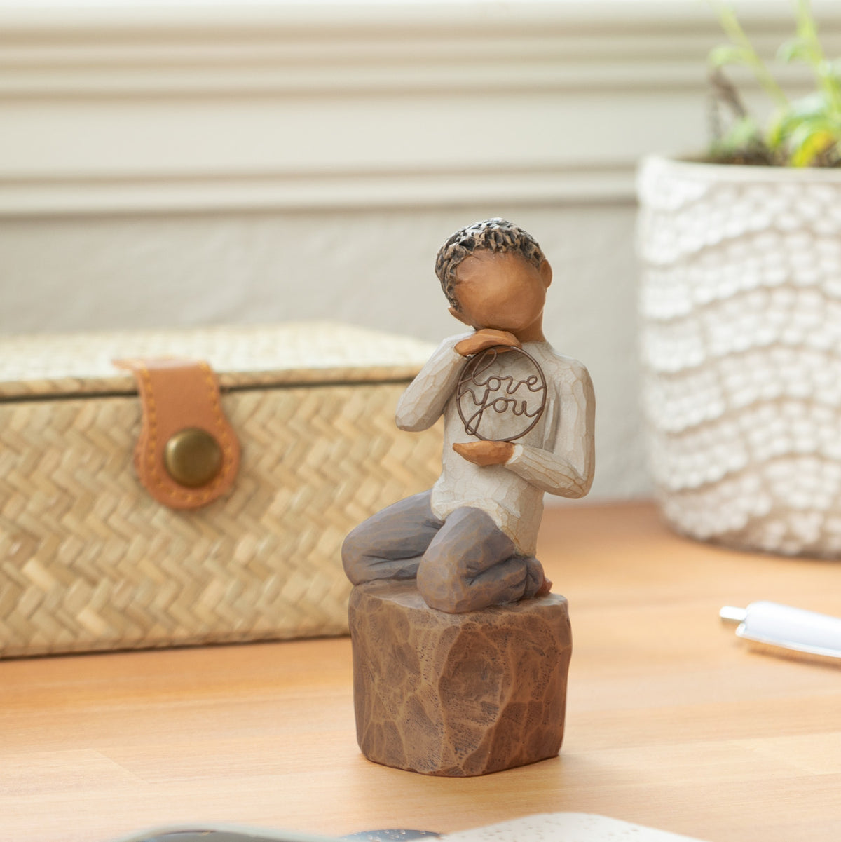 Willow Tree Love you too (darker skin), 4.5”h hand-painted resin figure of young child in cream shirt and jeans, kneeling on rock, holding wire circle with ‘love you’ inside, on desk