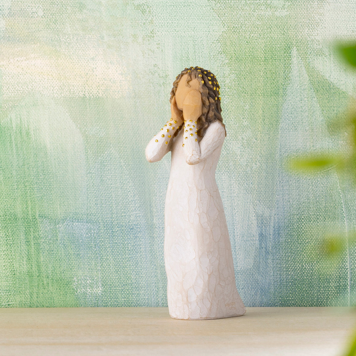 Willow Tree Littlest Christmas Angel, 5.5”h hand-painted standing resin figure in cream dress, both hands up to face in awe, gold leaf dots on sleeves and hair, in front of green painting
